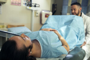 A person laying on a patient's seat while wearing a blue medical gown. There is a doctor looking at the person in the medical gown.