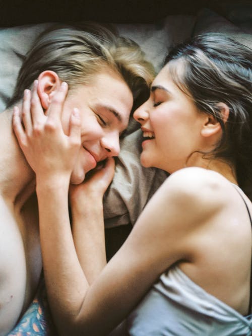 A couple laying in bed. They are smiling and one partner is holding the other person's face.