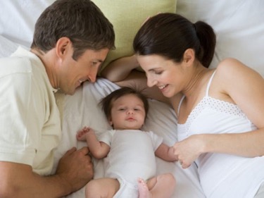 Man and woman laying in bed with their baby lying in the middle of them. Both look down smiling at their child. 