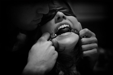 A blindfolded person with a ball gag in their mouth. Their partner is holding the straps of the ball gag.