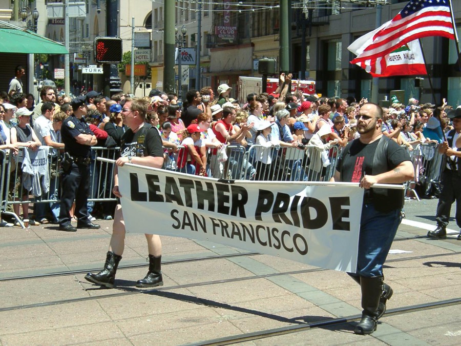 Two persons holding a banner that says "LEATHER PRIDE SAN FRANCISCO." There is a large group of people behind them.