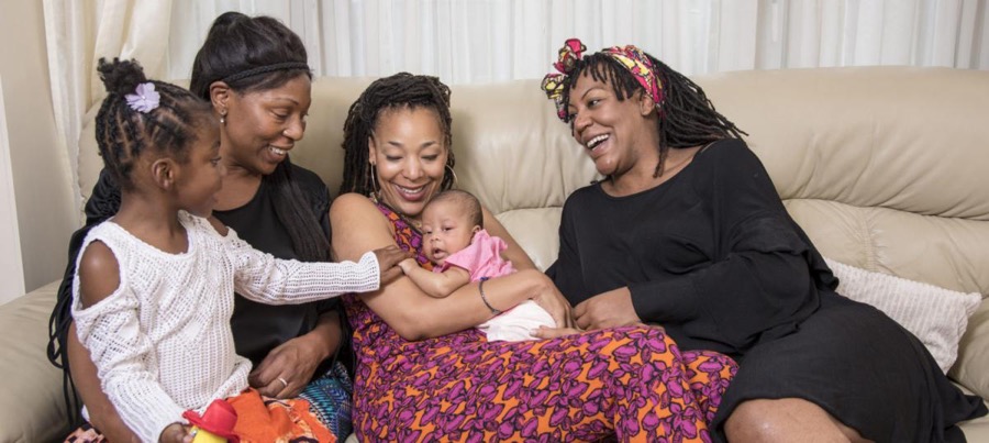 A family sitting laughing and smiling on a white couch together, consisting of three women, one young girl, and one new born