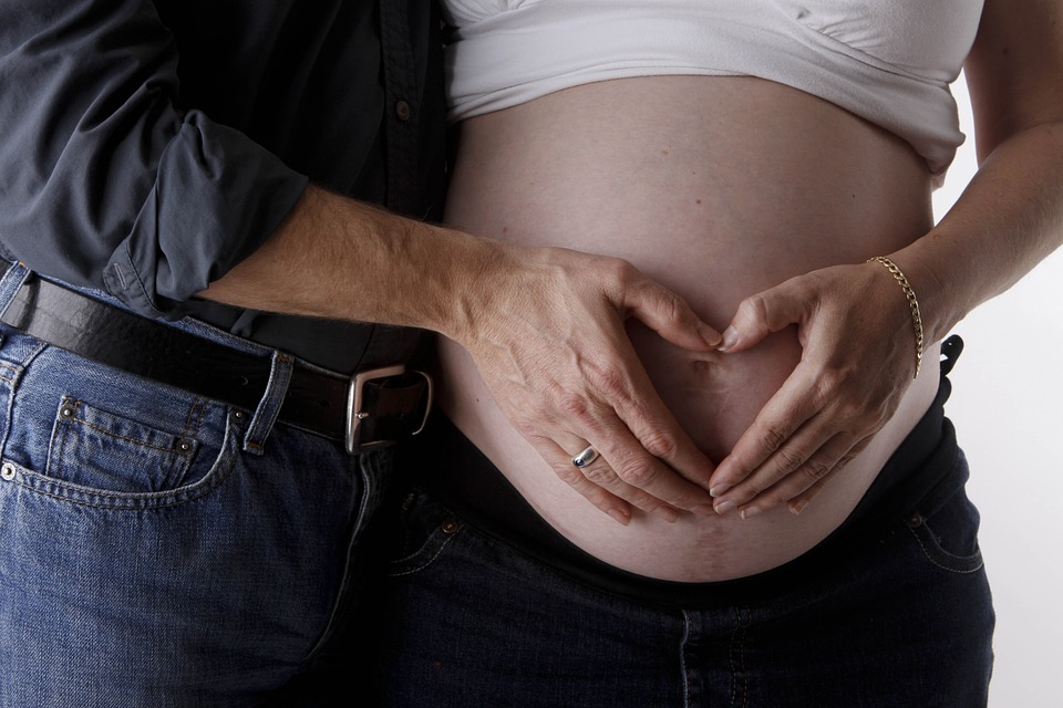 A person holding their partner's pregnant belly and forming a heart with their hands above the belly.