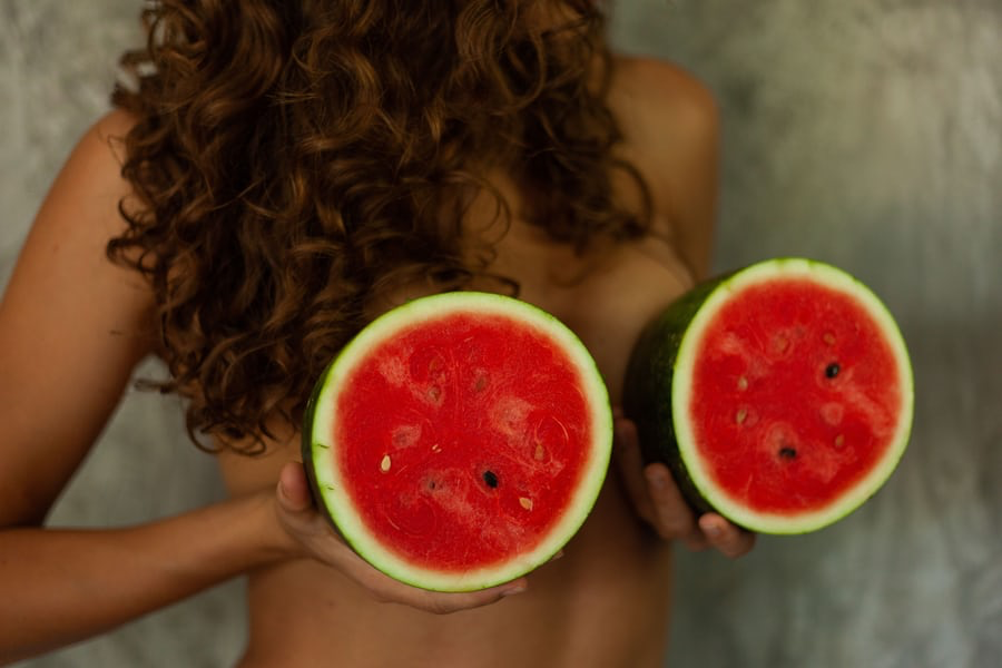 A shirtless woman holding two halves of a watermelon in front of each of her breasts