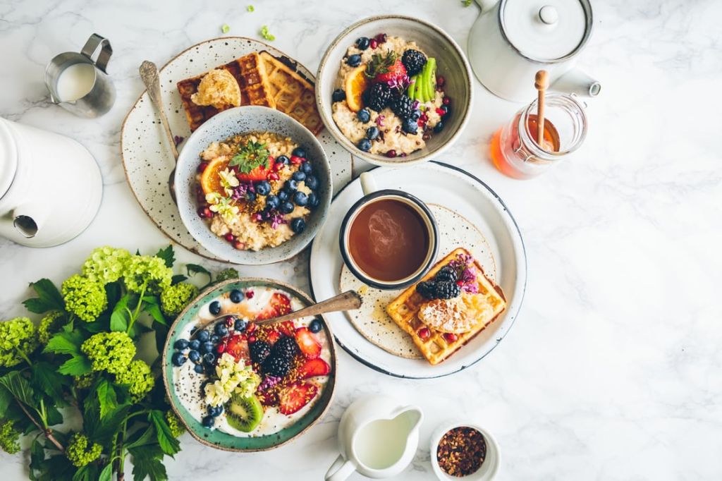 Bowl of fruit next to a plate of waffles, a cup of tea, a cup of milk, another bowl of fruit, a third bowl of fruit on top of a plate of two waffles, next to green flowers, all on a white marble table