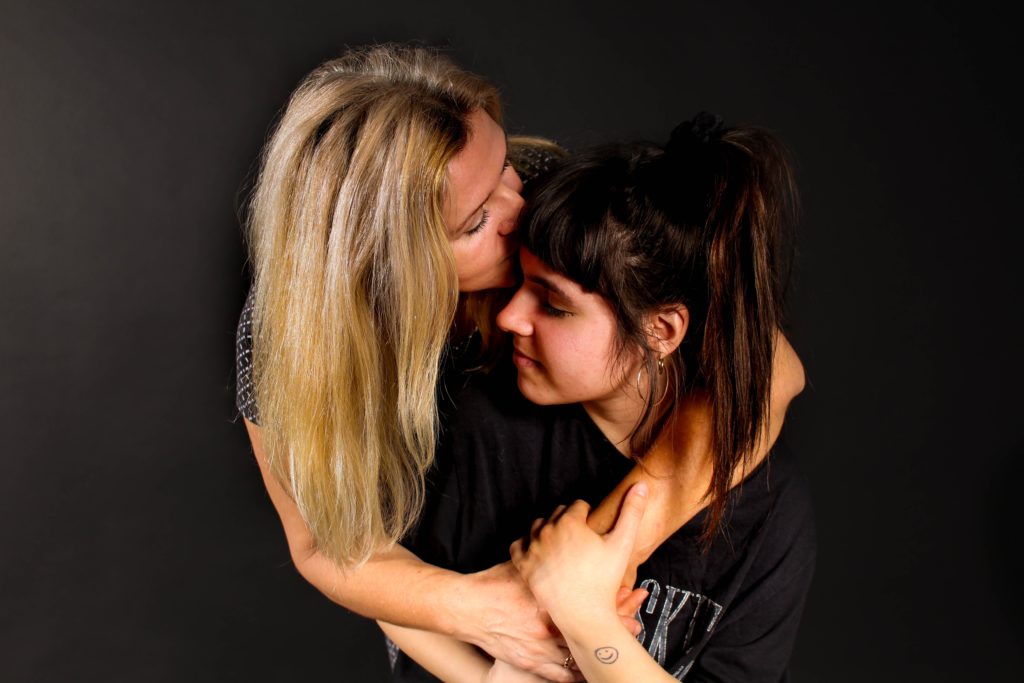 One blond woman with her arms around a brunette woman, kissing her forehead