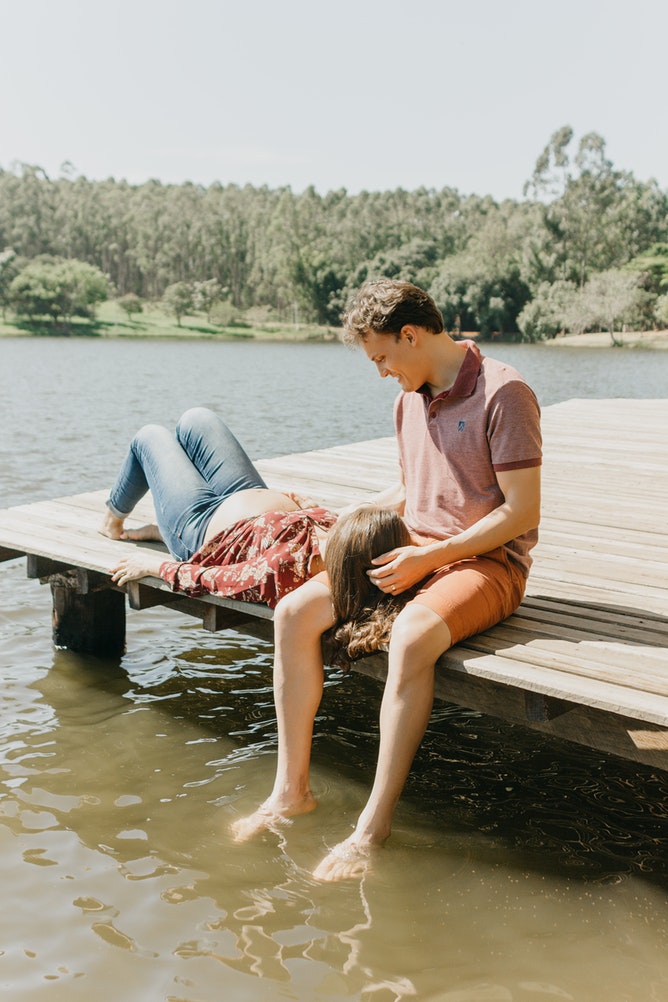 A couple sitting on a dock. The man has his feet in the water and the woman is laying her head on her partner's thighs.