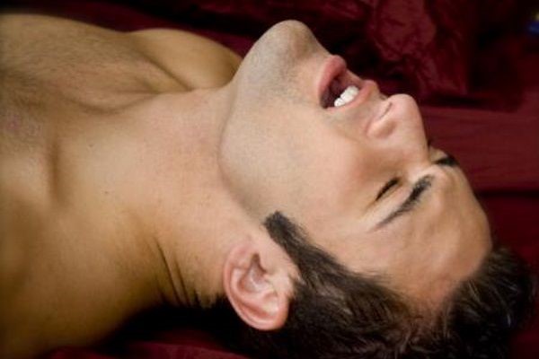 Shirtless man in bed, whose face indicates he is orgasming