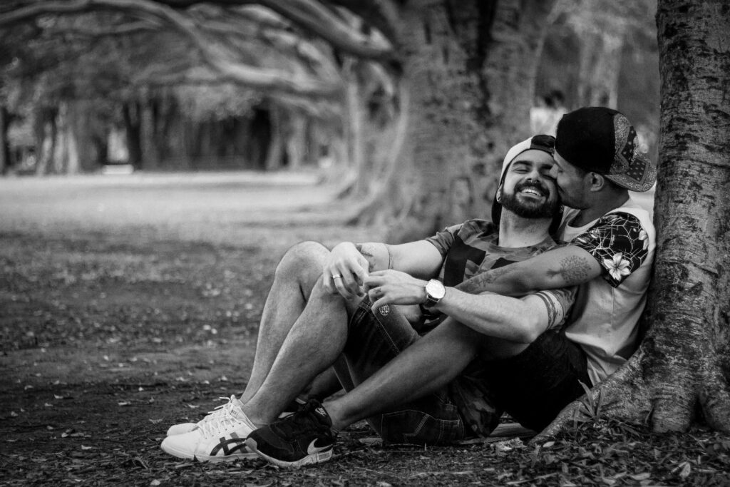 A man's arms wrapped around another man and kissing him, as the other smiles, by a tree