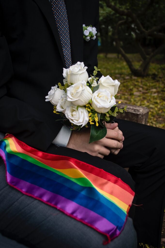 A person in a suit holding a bouquet of white roses and there is a rainbow flag next to their legs.