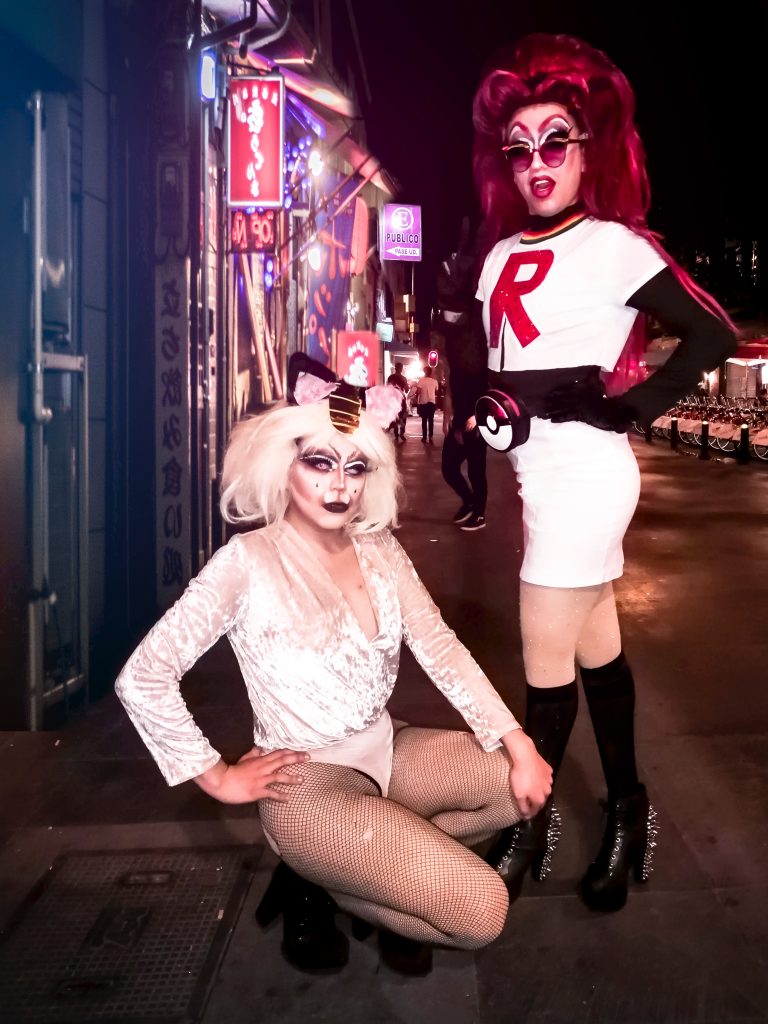 Two "drag queens" posing for the camera. Both are wearing heavy makeup and feminine clothing.