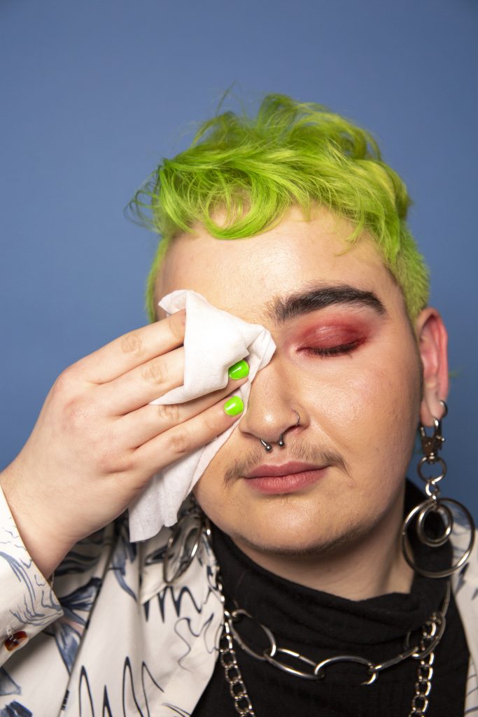 A person with short, green hair and green painted nails. The person is wiping their eye; they are wearing eyeshadow and jewelry.
