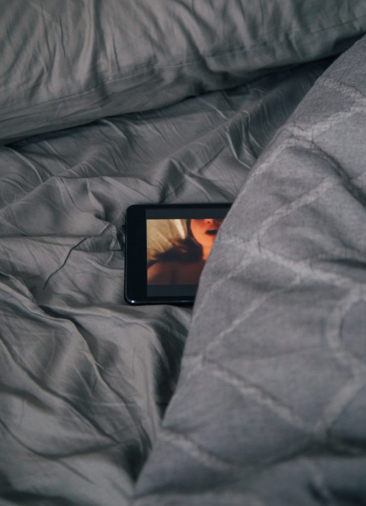 A phone showing a nude woman with her mouth open. The phone is in between bed sheets.