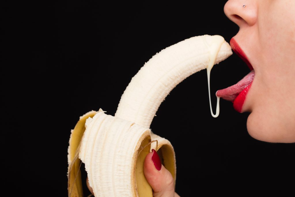 A person with their tounge sticking out and holding on to a peeled banana with a milky substance dripping.