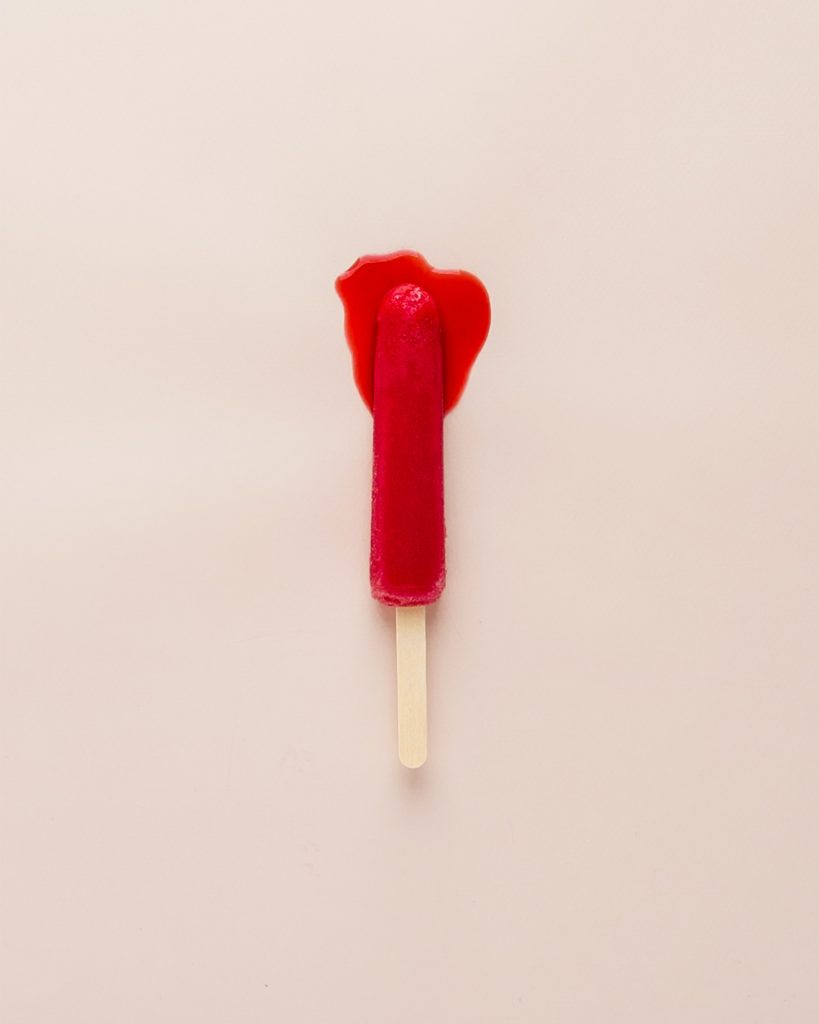 A red popsicle on a stick leaking red fluids; it resembles a bloody tampon.