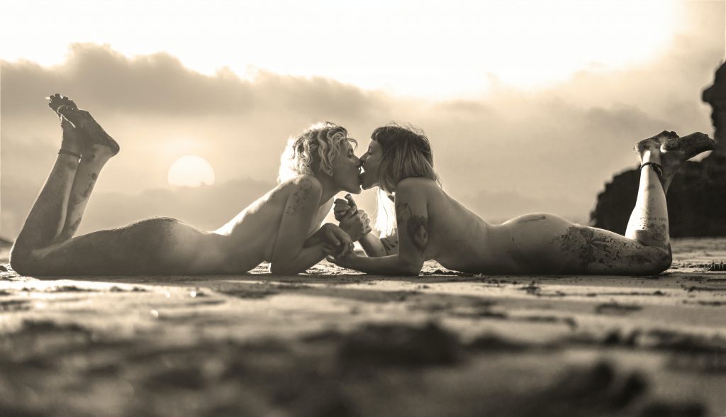 Two naked women lying down on the floor, facing each other, and tounge kissing.