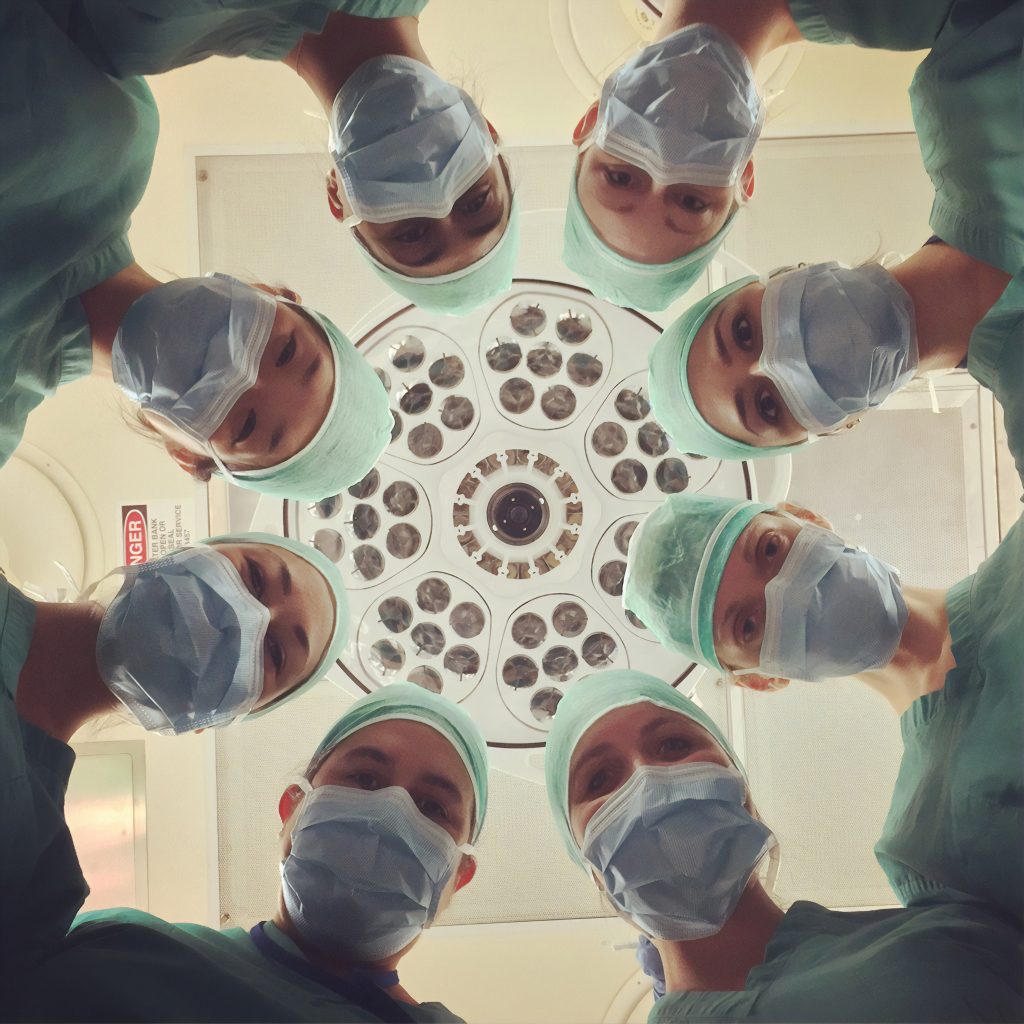 Medical professionals forming a circle with their heads. They are looking down and are wearing surgical masks and caps.