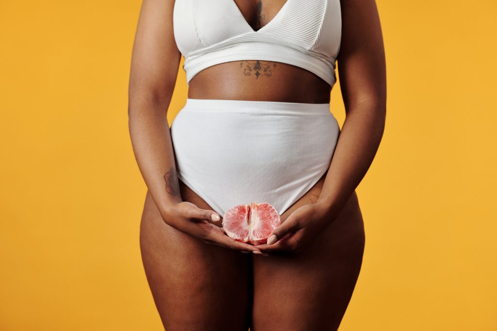 A person wearing a white bikini and holding half of a grapefruit that resembles a vagina. The grapefruit is hovering over her vulva area.