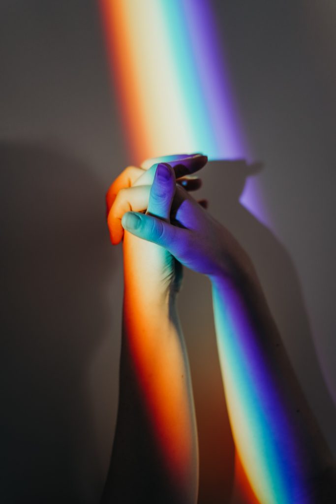 Two persons hands intertwined. There is a rainbow over the hands.