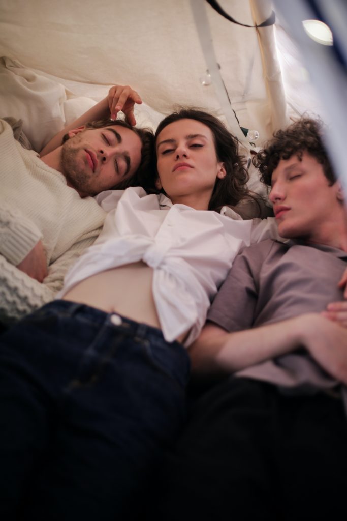 Three persons laying on a bed with their eyes closed. The person in the middle has their hands wrapped around the other two people.