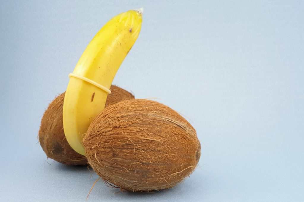 A banana wrapped in a condom and in between two kiwis. The banana resembles a penis and the kiwis resemble testicles.