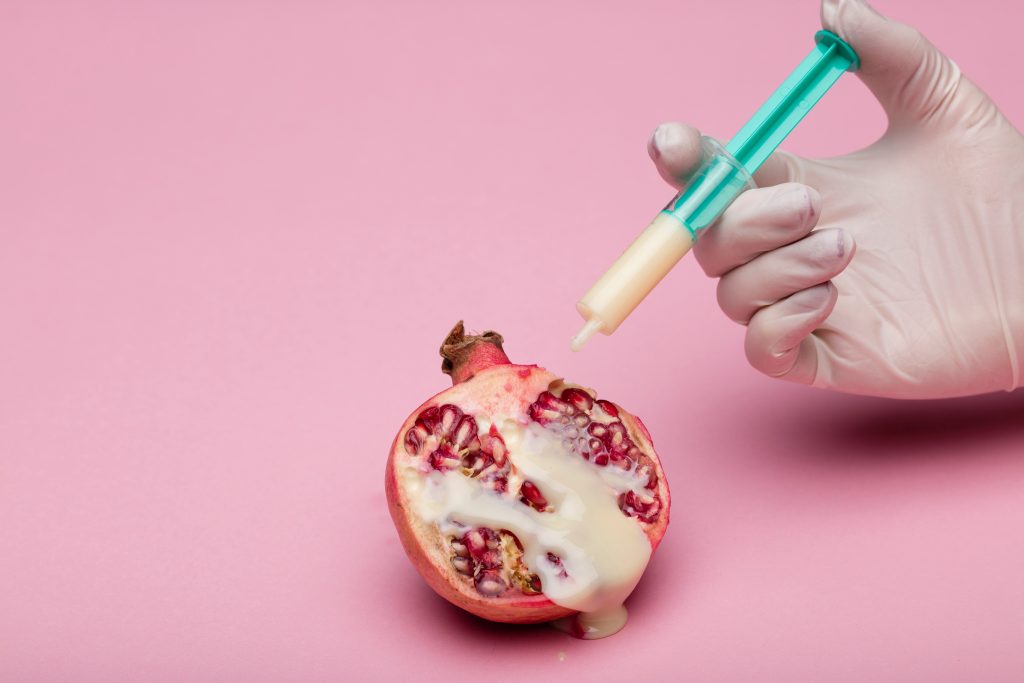 A hand holding a syringe filled with a milky substance. The milky substance is dripping on half of a pomegranate.