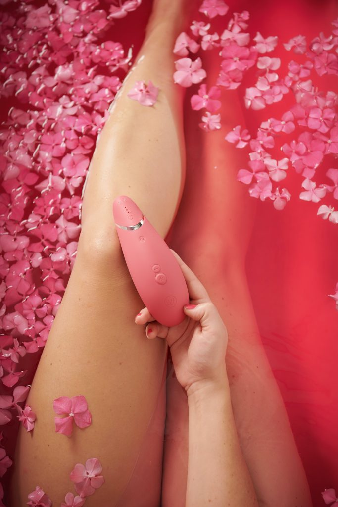 A person's legs in a bathtub filled with pink water and pink flowers. The person is holding a pink vibrator.