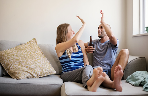 A couple sitting on a couch, both man and woman are smiling and reaching up to press their hands together in a high-five.