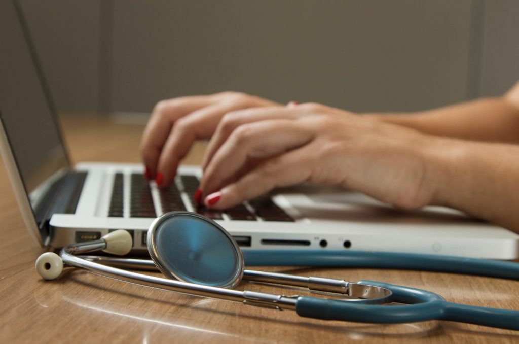 Person's hands typing on laptop computer adjacent to a stethoscope.