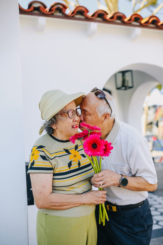 An older couple standing and holding a bouquet of flowers together. The man is pressing a kiss to the woman’s cheek. 