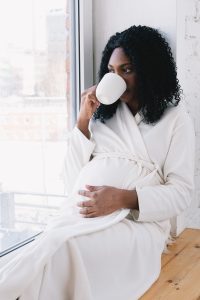 Black woman drinking beverage and clutching pregnant belly. 