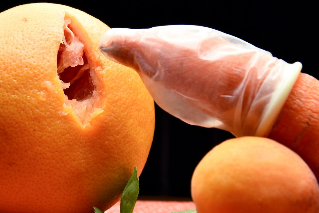 A carrot wearing a condom entering an orange with a small hole.