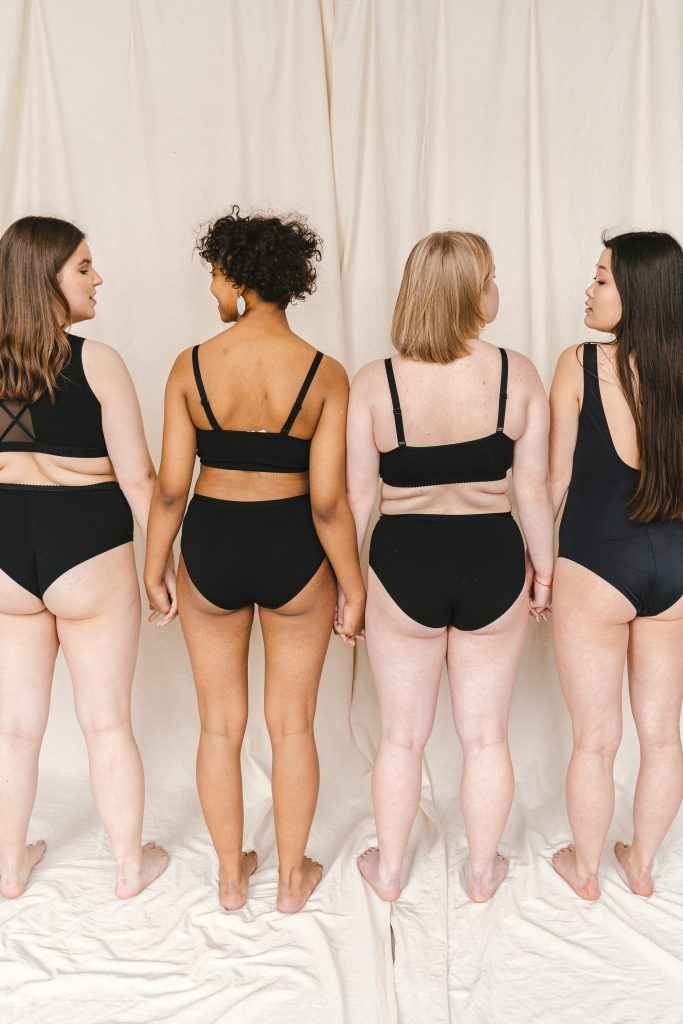Four women wearing black swimsuits; they are all holding hands and facing a white curtain.