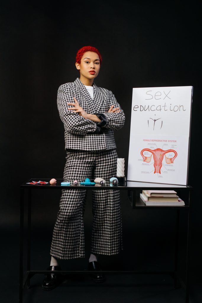 A person standing next to a poster with "Sex education" written on it, as well as a diagram of the female reproductive system. In front of the person, there is a variety of sex toys, condoms, and books.