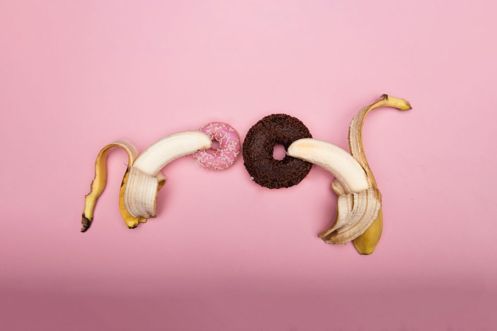 Two peeled bananas on top of two donuts. One donut is small and pink. The other donut is bigger and brown.
