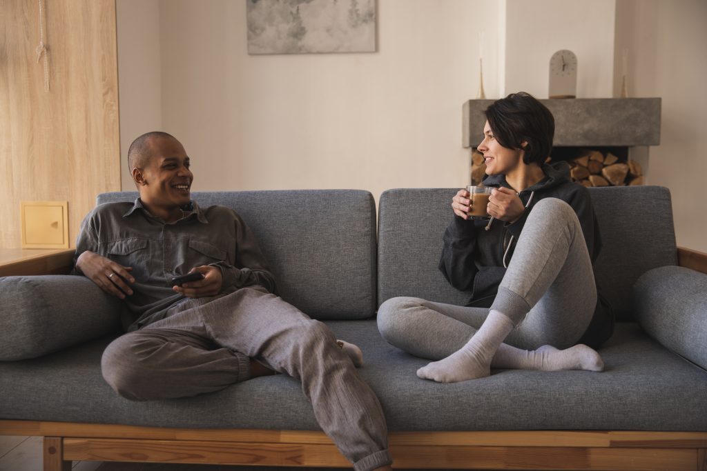 Man and woman sit across from each other on a grey couch while smiling and enjoying conversation. 