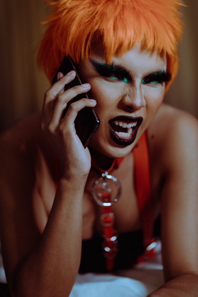 A person with orange hair, dark lipstick, and dramatic makeup holding a phone to their ear.