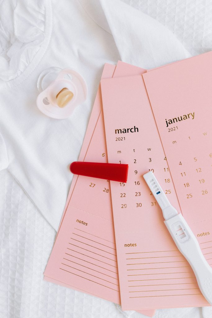 A pregnancy test on top of pink monthly calendar and next to a baby pacifier.