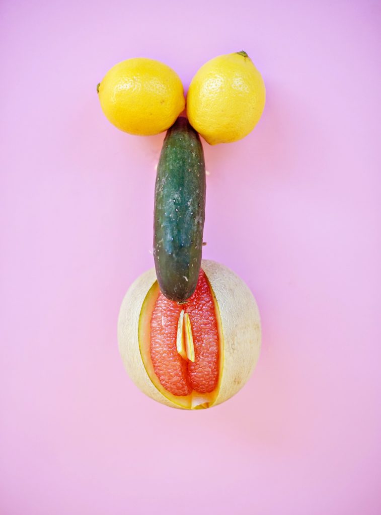 A lemon on each side of a cucumber. The cucumber is on top of a melon that is shaped like a vulva.