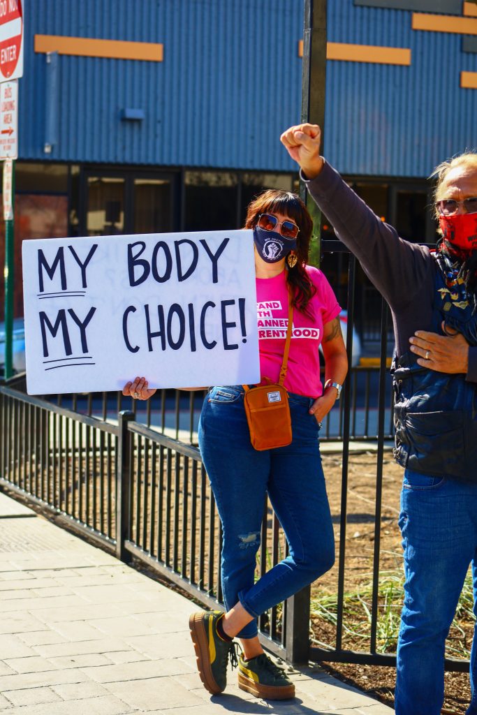 A person wearing a shirt that says "I STAND WITH PLANNED PARENTHOOD" and the person is holding a poster that says "MY BODY MY CHOICE!"