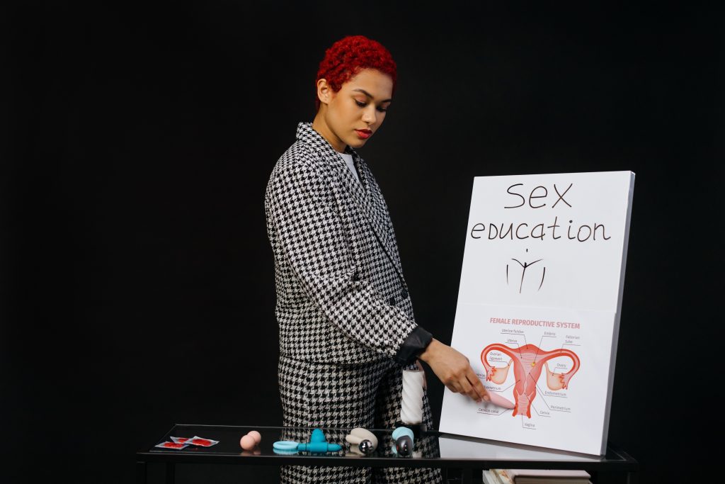 A person standing next to a poster that says "sex education" and has a diagram of the female reproductive system. There is a table with vibrators and condoms in front of the person.