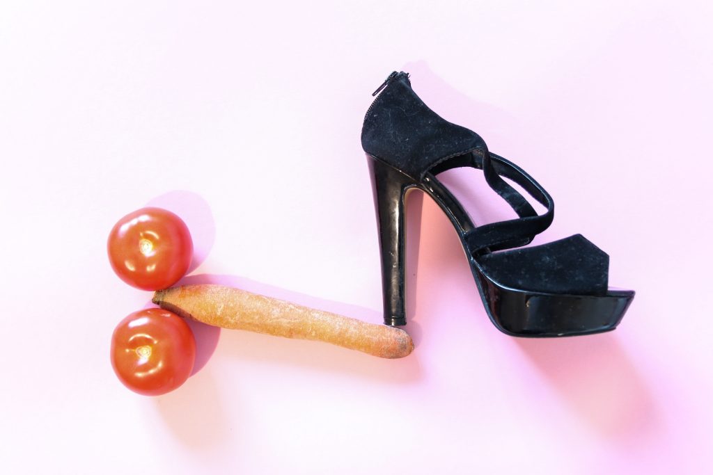 A black high heel over a carrot with a tomato on each of its side, shaped like a penis.