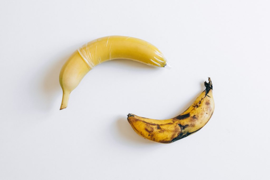 A yellow banana with a condom next to a very ripe, spotty banana.