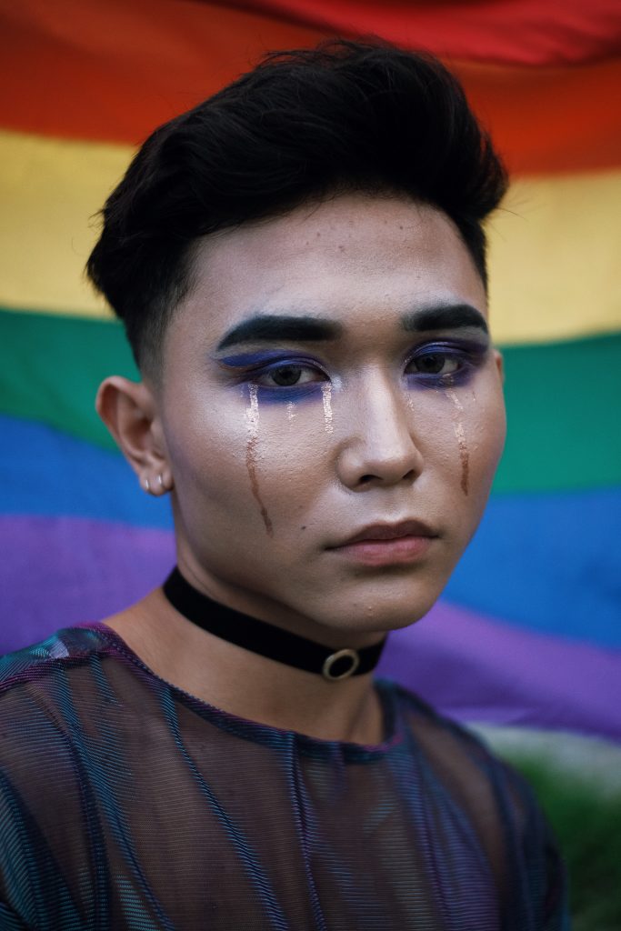 A close-up of a person wearing makeup. They are standing in front of a rainbow flag.