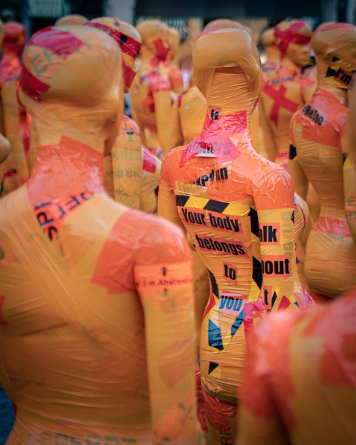 Several yellow and orange mannequins in a crowd, with text pasted on them that reads: "Your body belongs to you".