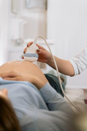 A doctor wears an ultrasound transducer across the pregnant patient's belly. The patient lies down on their back with their belly exposed.