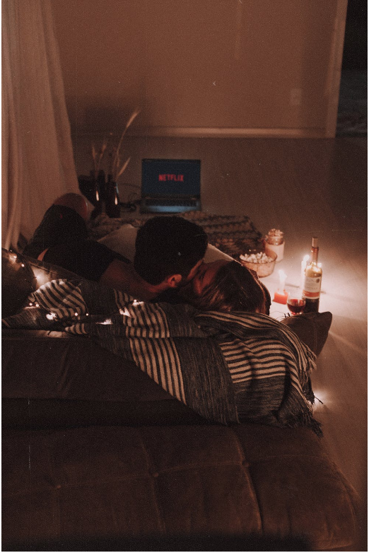 Couple cuddling on a floor cushion with netflix loading screen on the laptop in front of them. There is also popcorn and a bottle of wine on the floor next to the couple. 