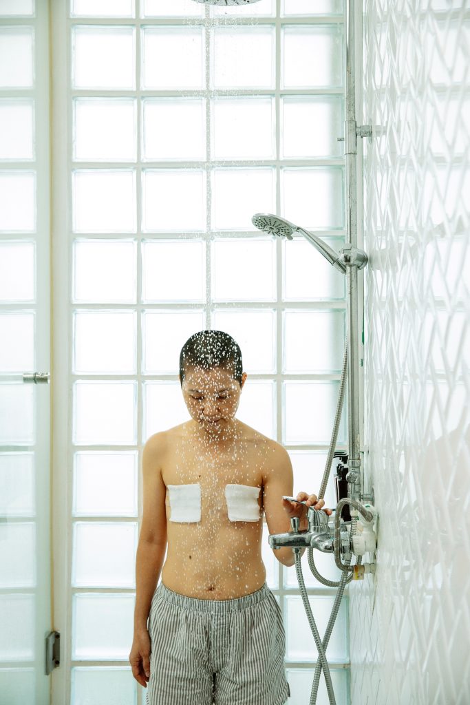 A person with bandages on their chest from mastectomy is in the shower. They are looking down.
