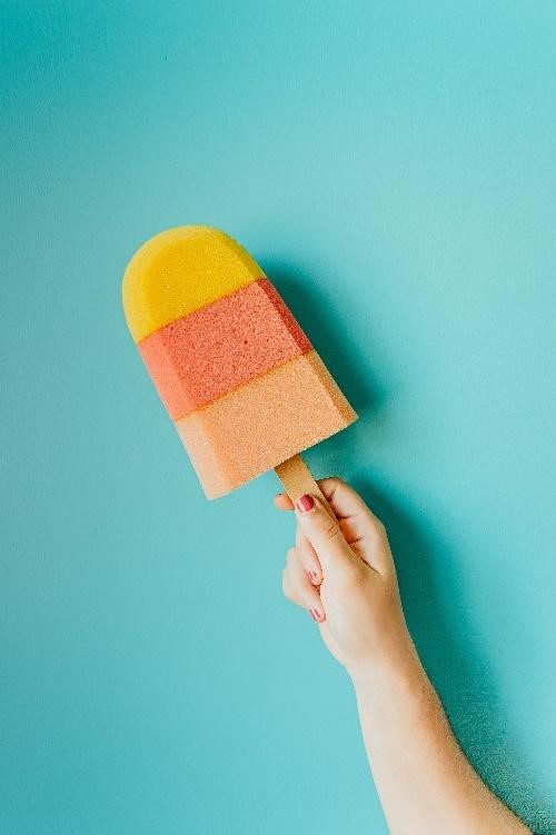 A hand holding a sponge shaped like a popsicles over a blue background.