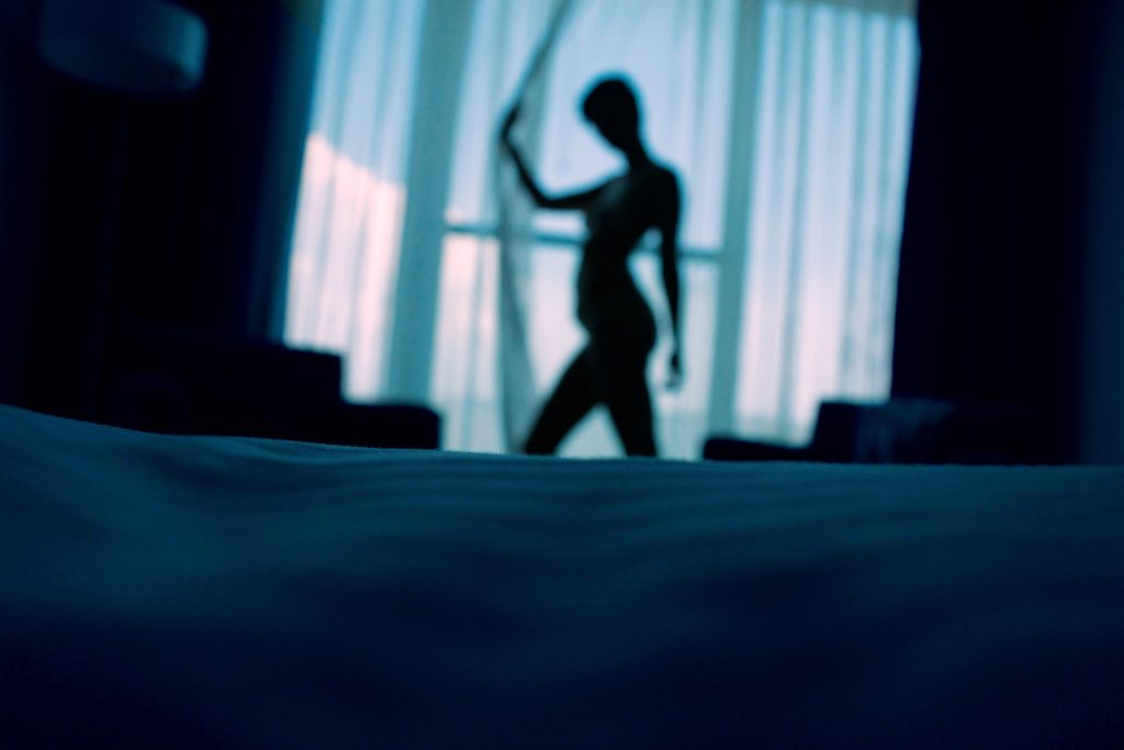 The silhouette of a nude person holding on to a window curtain.
